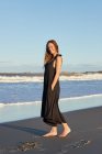 Vue latérale de la femme souriante en robe d'été debout sur le bord de mer sablonneux et regardant la caméra — Photo de stock
