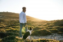 Corpo inteiro visão traseira da mulher étnica feliz com Border Collie cão andando juntos na trilha entre colinas gramadas na noite de primavera ensolarada — Fotografia de Stock
