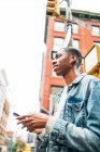 From below cheerful content African American guy in denim trendy jacket surfing modern mobile phone during city stroll — Stock Photo