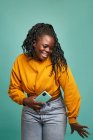 Sorrindo afro-americano feminino em jeans e camisola amarela segurando smartphone moderno e dançando contra a parede azul em estúdio — Fotografia de Stock