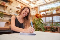 Niedriger Winkel der fröhlichen Gärtnerin sitzt am Tisch und schreibt in Notizblock im Gewächshaus — Stockfoto