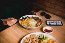 Frau in lässiger Kleidung mit Smartphone und Sonnenbrille isst leckeren taiwanesischen Bento mit Essstäbchen in asiatischem Café — Stockfoto