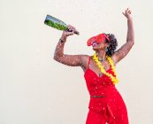 Fröhliche Afroamerikanerin in roter Kleidung und Brille mit einer Flasche alkoholischen Getränks während einer Party auf hellem Hintergrund — Stockfoto
