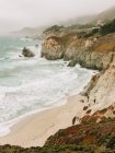 Angle élevé de merveilleux paysages d'eau de mer orageuse avec des vagues de mousse qui lavent la plage de sable contre le rivage brumeux des hautes terres sous un ciel gris à Big Sur — Photo de stock