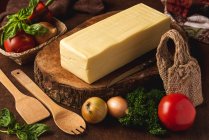 Блок сиру на дерев'яній підставці біля сирої цибулі і тканий мішок проти органічних шпателів і листя базиліка — стокове фото