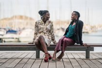 Стильные улыбающиеся афроамериканки проводят время вместе, сидя на деревянной скамейке в парке в яркий день — стоковое фото