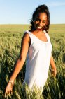 Lächelnde junge schwarze Dame in weißem Sommerkleid schlendert auf grünem Weizenfeld, während sie tagsüber unter blauem Himmel in die Kamera schaut — Stockfoto