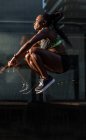 Vista lateral de una mujer negra fuerte saltando alto cerca de la pared de vidrio del edificio moderno mientras hace ejercicio en la calle de la ciudad en un día soleado - foto de stock