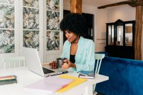 Joven freelancer afroamericana en traje casual bebiendo café mientras está sentada en la mesa y trabajando en el proyecto con el portátil en casa - foto de stock