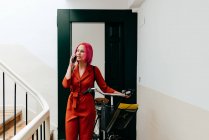 Молода стильна жінка в червоному костюмі з рюкзаком розмовляє на смартфоні, стоячи з велосипедом на сходах — стокове фото