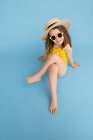 Высокий угол полный тела милой счастливой маленькой девочки в желтом купальнике и соломенной шляпе со стильными солнцезащитными очками сидя на синем фоне и глядя в камеру — стоковое фото