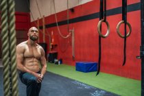 Hombre barbudo muscular mirando hacia arriba mientras está de pie cerca del equipo durante el entrenamiento en el gimnasio moderno - foto de stock