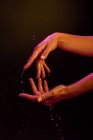Vista ritagliare di donna anonima facendo gesti artistici con le mani sotto le luci rosa e gialle e spruzzi d'acqua sullo sfondo nero — Foto stock