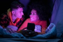 Glückliche multiethnische Männer und Frauen lächeln und schauen einander an, während sie sich ausruhen und nachts im Zelt nach dem Handy suchen — Stockfoto