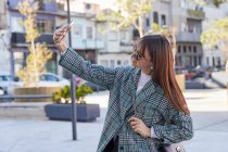 Молода стильна жінка в модному пальто і сонцезахисні окуляри беруть селфі на смартфон, стоячи на міській вулиці — стокове фото