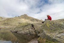 Male photographer with camera in outerwear sitting on stony coast near tranquil water of Laguna Grande lake against cloudy sky in Circo de Gredos cirque in Avila, Spain — Stock Photo