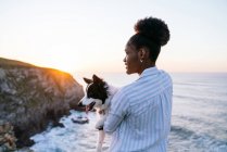 Vista lateral do proprietário feminino afro-americano segurando bonito feliz Border Collie cão enquanto desfruta de tempo juntos perto do mar acenando ao pôr do sol — Fotografia de Stock