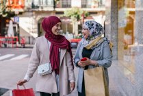 Веселі мусульманські друзі-жінки з паперовими мішками, що ходять по місту після покупки, дивлячись один на одного — стокове фото