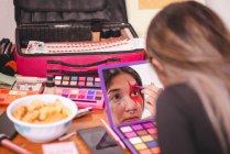 Joven mujer mirando el espejo y la pintura ornamento en la cara mientras se aplica maquillaje creativo en el estudio - foto de stock