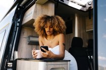 Glad African American woman with mug of hot beverage smiling and browsing cellphone while resting in contemporary caravan in morning — Stock Photo