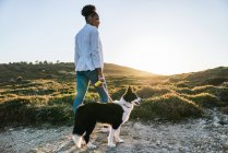 Corpo pieno vista posteriore di felice donna etnica con Border Collie cane camminare insieme sul sentiero tra le colline erbose in soleggiata serata primaverile — Foto stock