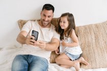 Pai alegre e menina sentados na cama e tomando auto-retrato no smartphone enquanto se divertem juntos durante o fim de semana em casa — Fotografia de Stock