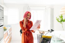 Joven diseñadora creativa en traje de moda y anteojos sosteniendo papel con plantillas y hablando en smartphone mientras está de pie en un moderno apartamento con luz - foto de stock