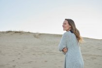 Содержание женщины в теплой одежде, стоящей на пляже у моря и наслаждающейся летним вечером, глядя в сторону — стоковое фото