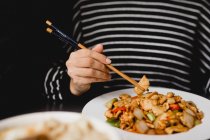 Femme méconnaissable utilisant des baguettes pour manger une portion de délicieux poulet Gong Bao sur fond noir au restaurant — Photo de stock