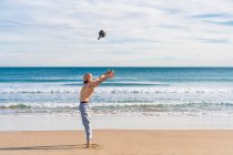 Vue latérale de l'homme sportif sans chemise jetant kettlebell sur la côte sablonneuse avec des vagues de l'océan sur fond — Photo de stock