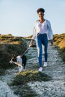 Ganzkörper glückliche ethnische Frau mit Border Collie Hund beim gemeinsamen Spaziergang auf dem Weg zwischen grasbewachsenen Hügeln an sonnigen Frühlingsabend — Stockfoto