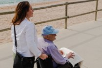 Encantada filha adulta empurrando cadeira de rodas com pai sênior e curtindo passeio ao longo do passeio perto do mar — Fotografia de Stock