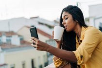 Молодая латиноамериканка наслаждается свежим кофе и пользуется мобильным телефоном, расслабляясь на балконе вечером — стоковое фото