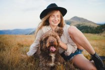 Cheerful female owner with obedient Labradoodle dog sitting in mountains looking away — Stock Photo