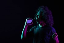 Unbekümmerter Latin-Männchen mit Vintage-Kassettenspieler und Kopfhörer, der Musik hört und Lieder auf schwarzem Hintergrund im Studio mit Neonlicht singt — Stockfoto