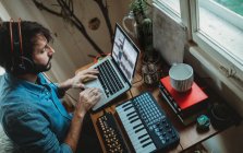 Сверху вид сфокусированного молодого человека в наушниках, работающего на синтезаторе и ноутбуке дома — стоковое фото
