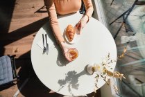 Mujer anónima en boina sentada a la mesa en la cafetería con vaso aromático de café y croissant recién horneado - foto de stock