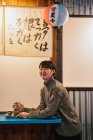 Vista lateral de la mujer asiática muy alegre en suéter sentado en la mesa azul con bebida tradicional en la cafetería - foto de stock
