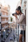 Vista lateral étnica femenina en desgaste con adorno a rayas con dispositivo fotográfico profesional mirando hacia otro lado en el balcón durante el día - foto de stock