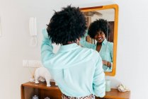 Back view of a beautiful young African American female in casual outfit touching curly hair while standing in room and looking at mirror — Stock Photo