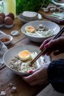 Cropped unrecognizable person preparing fresh cooked ramen noodles with tofu, eggs and vegetables with chopsticks on a wooden table — Stock Photo