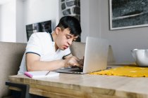 Enfoque adolescente latino con síndrome de Down netbook de navegación mientras está sentado en la mesa y estudiar en línea desde casa - foto de stock