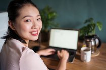 Бічний вид щасливої азіатської жінки-фрилансер читає документи на ноутбуці, сидячи за столом під час віддаленої роботи, дивлячись на камеру. — стокове фото