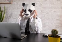 Empresario masculino cansado anónimo con máscara de oso panda y camisa blanca trabajando en la mesa con netbook en el espacio de trabajo - foto de stock