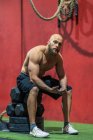 Vista lateral do homem musculoso exausto olhando para a câmera sentada em pesos e descansando durante o treino funcional no ginásio — Fotografia de Stock
