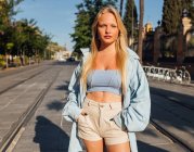 Charming female with blond hair and in trendy summer clothes standing with hands in pockets in city and looking at camera — Stock Photo