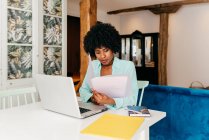 Young African American female freelancer in casual outfit drinking coffee while sitting at table and working on project with laptop at home — Stock Photo