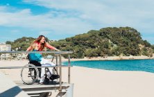 Вид сбоку на взрослую женщину с пожилой матерью в инвалидной коляске, любовающуюся видом на море в солнечный день — стоковое фото
