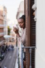 Vista laterale della donna afroamericana riflettente con tazza di bevanda calda guardando la fotocamera dal balcone in città — Foto stock