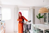Young creative female designer in trendy outfit and eyeglasses talking on smartphone while standing with bicycle in modern light apartment — Stock Photo